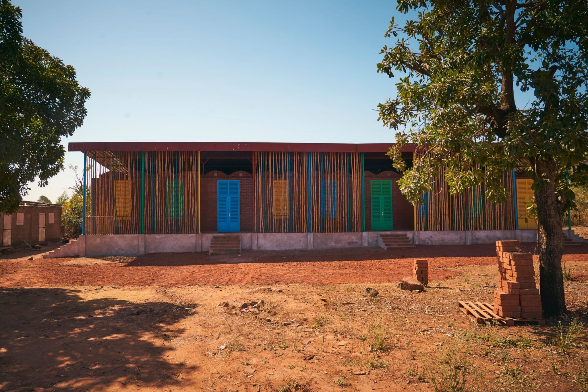École Kalan Deme So à Dougourakoro : bâtiment de classes bas, bardage de bambous colorés, entre deux karités, au pied d'une colline rocheuse.